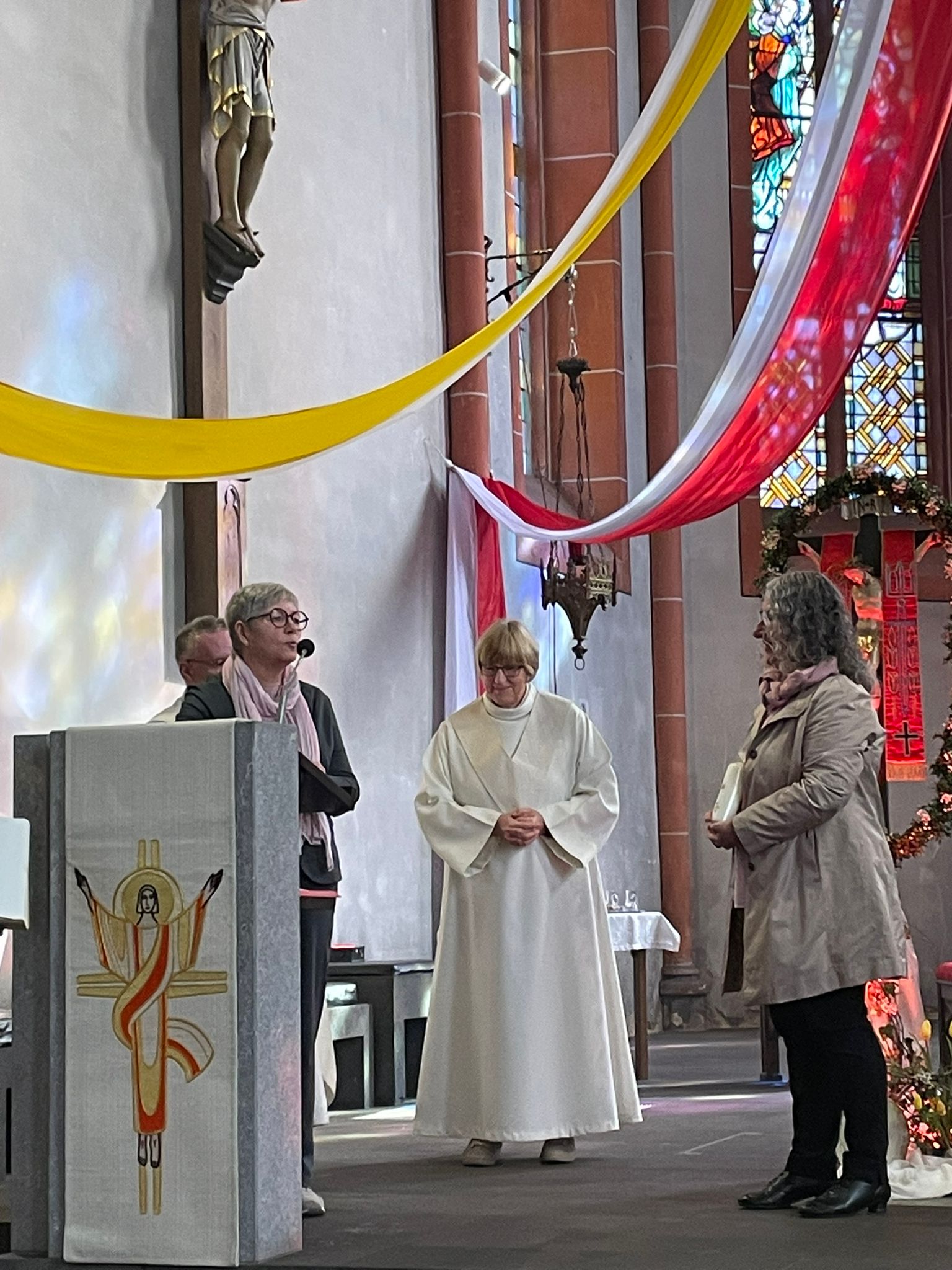 eine Frau mit einer weißen Albe unzwei Frauen im Altarraum einer festlich geschmückten Kirche, ein Priester steht im Hintergrund