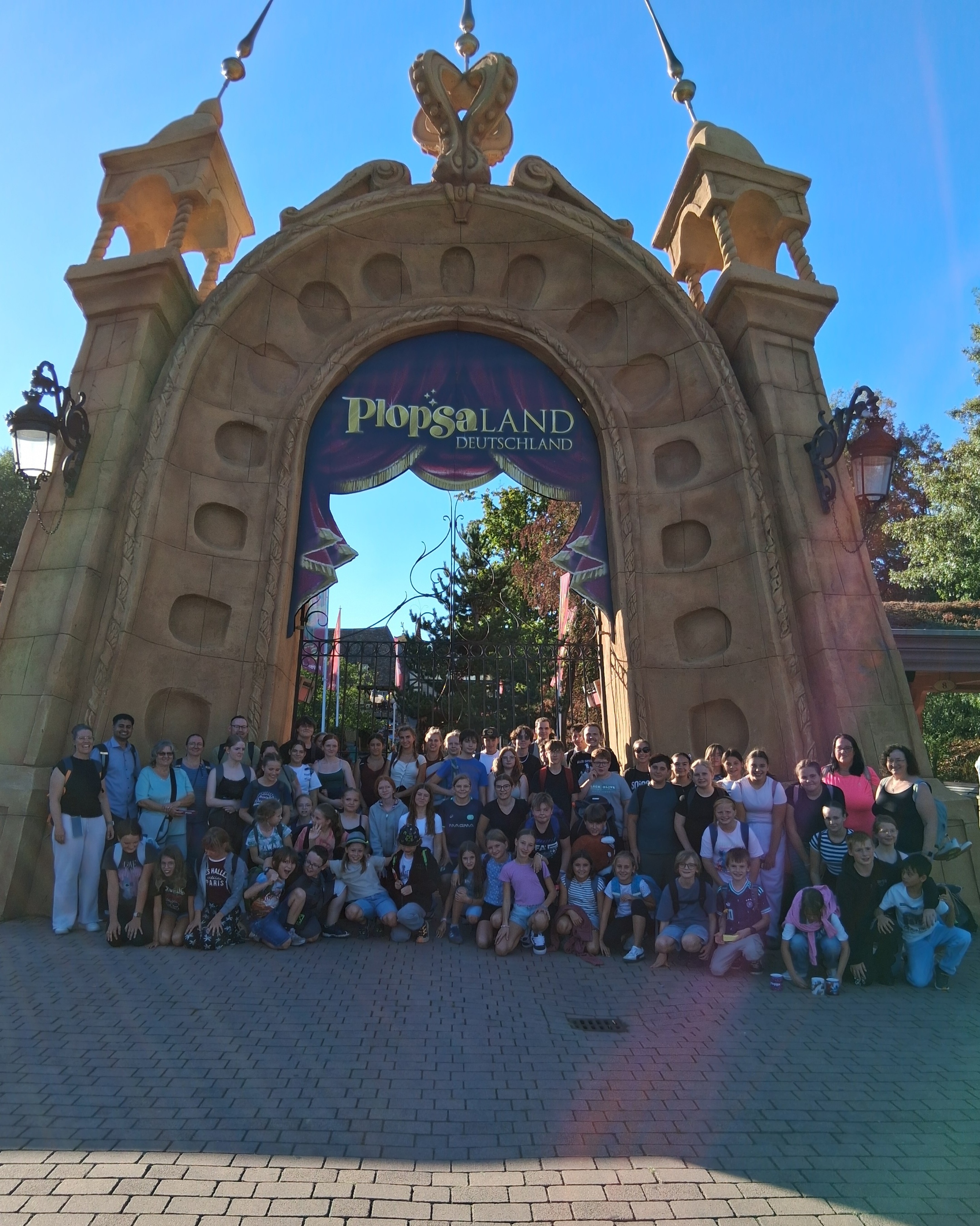 Gruppenfoto von rund 70 Kindern, Jugendlichen und Begleitpersonen vor dem großen, märchenhaft gestalteten Eingangstor des Freizeitparks Plopsaland Deutschland. Über dem Tor befindet sich die Aufschrift ‚Plopsaland Deutschland‘. Die Gruppe steht und kniet auf einem gepflasterten Platz vor dem Eingang. Darüber spannt sich ein sonniger, wolkenloser blauer Himmel. (c) Messdienerbetreuer PastR Lebach Gruppenfoto von rund 70 Kindern, Jugendlichen und Begleitpersonen vor dem großen, märchenhaft gestalteten Eingangstor des Freizeitparks Plopsaland Deutschland. Über dem Tor befindet sich die Aufschrift ‚Plopsaland Deutschland‘. Die Gruppe steht und kniet auf einem gepflasterten Platz vor dem Eingang. Darüber spannt sich ein sonniger, wolkenloser blauer Himmel.