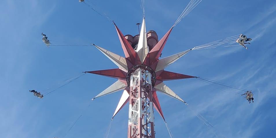 Ein hohes Kettenkarussell ragt vor einem strahlend blauen Himmel in die Höhe. Die Fahrgäste schwingen in ihren Sitzen an langen Ketten kreisförmig um den rot-weiß gestalteten Turm. Die Aufnahme wurde aus der Untersicht gemacht und betont die Höhe der Attraktion (c) Messdienerbetreuer PastR Lebach Ein hohes Kettenkarussell ragt vor einem strahlend blauen Himmel in die Höhe. Die Fahrgäste schwingen in ihren Sitzen an langen Ketten kreisförmig um den rot-weiß gestalteten Turm. Die Aufnahme wurde aus der Untersicht gemacht und betont die Höhe der Attraktion