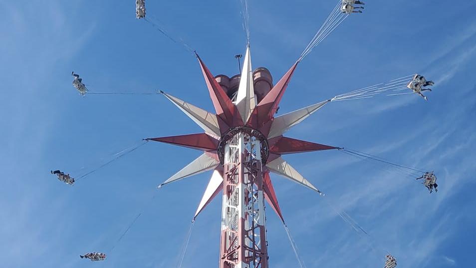 Ein hohes Kettenkarussell ragt vor einem strahlend blauen Himmel in die Höhe. Die Fahrgäste schwingen in ihren Sitzen an langen Ketten kreisförmig um den rot-weiß gestalteten Turm. Die Aufnahme wurde aus der Untersicht gemacht und betont die Höhe der Attraktion