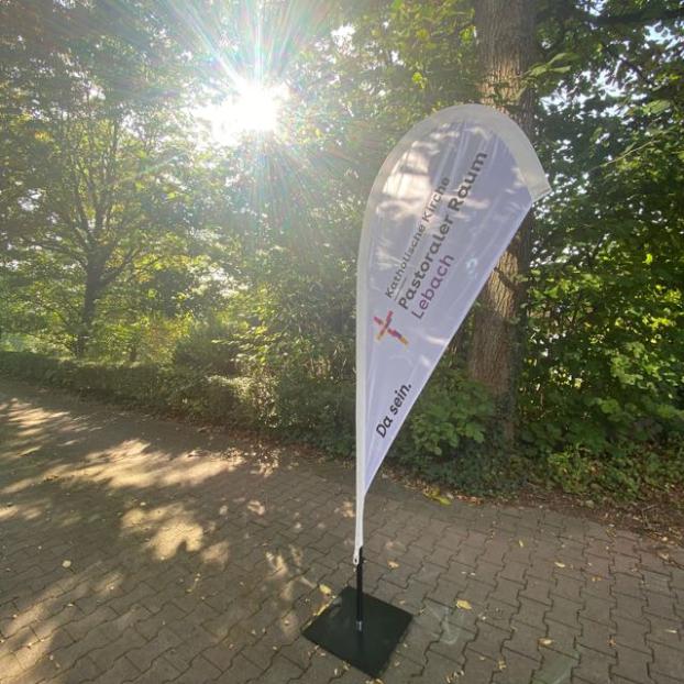 Eine schmale gepflasterte Straße führt durch einen grünen, schattigen Bereich mit vielen Bäumen und Sträuchern. Rechts im Vordergrund steht eine hohe, weiße, tropfenförmige Werbefahne auf einem schwarzen Fuß. Auf der Fahne steht gut sichtbar der Text „Katholische Kirche Pastoraler Raum Lebach“ sowie der Slogan „Da sein.“ und ein kleines Kreuzsymbol. Die Sonne scheint von links durch die Baumkronen und erzeugt helle Lichtstrahlen und starke Lichtreflexe. Der Boden ist teilweise im Schatten, teilweise von Sonnenlicht beleuchtet. Im Hintergrund sind dichte Büsche und weitere Bäume zu sehen, die eine ruhige, natürliche Umgebung bilden.